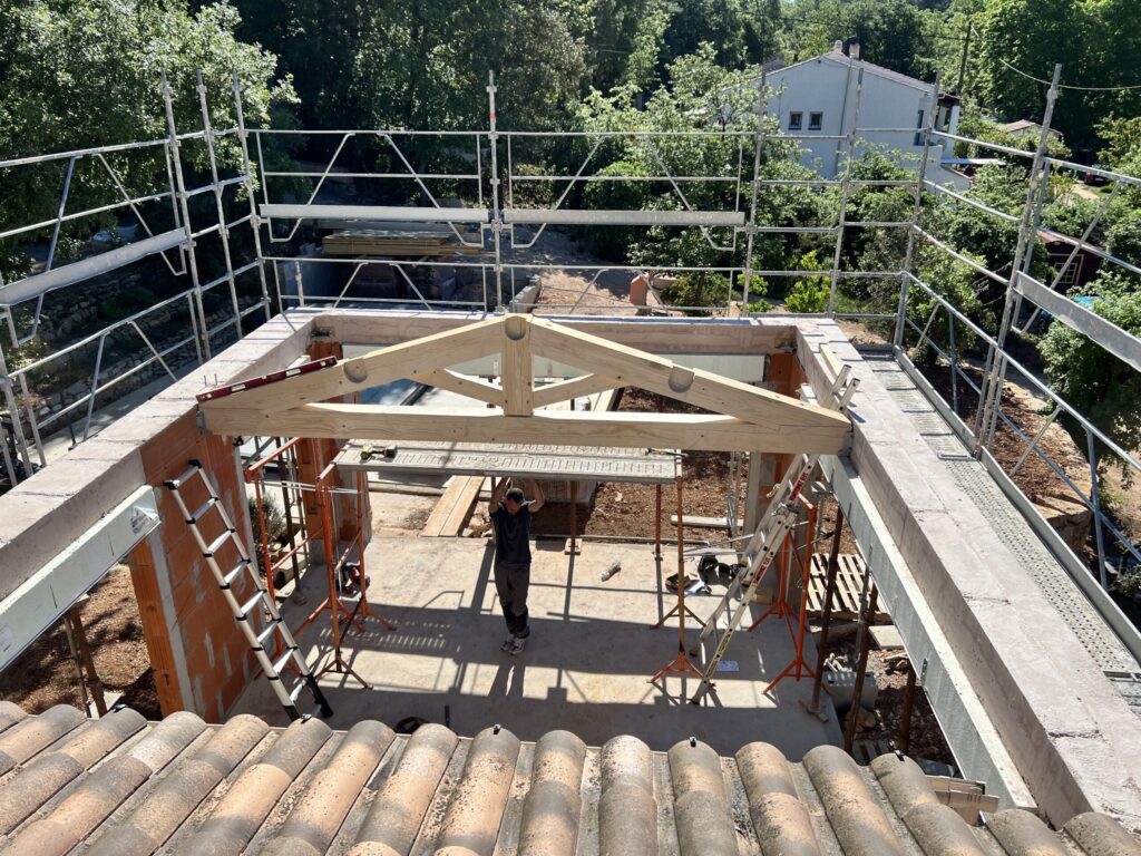 Chantier de construction d'une maison avec charpente en bois, échafaudages métalliques et ouvrier sur site, entouré de